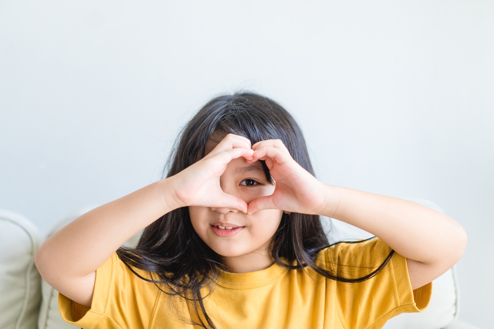 A young girl with a radiant smile, playfully framing her eyes with her hands formed into a heart shape