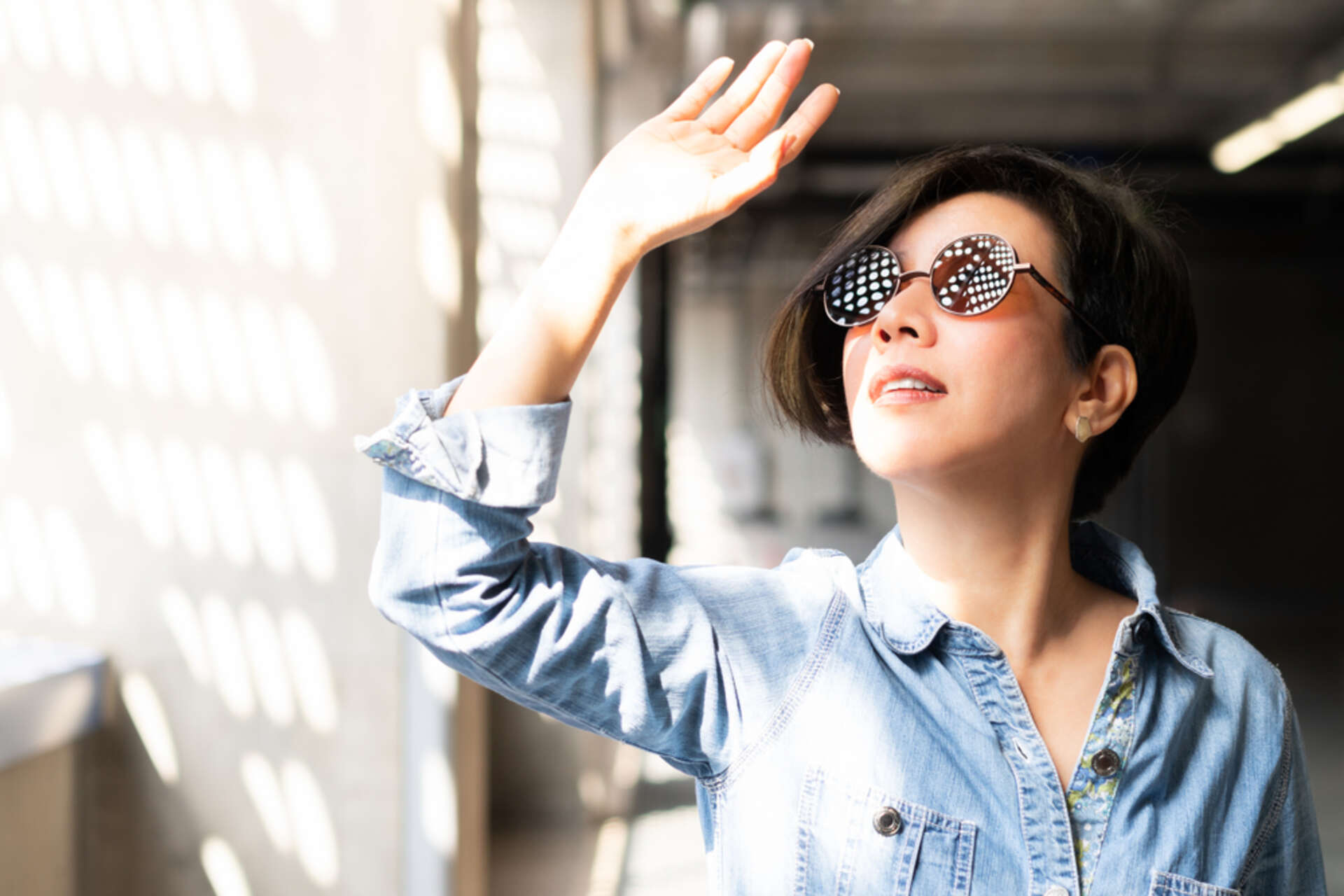 A woman in stylish sunglasses and a denim shirt shields her eyes with her hand from bright sunlight, symbolizing the importance of summer eye care.