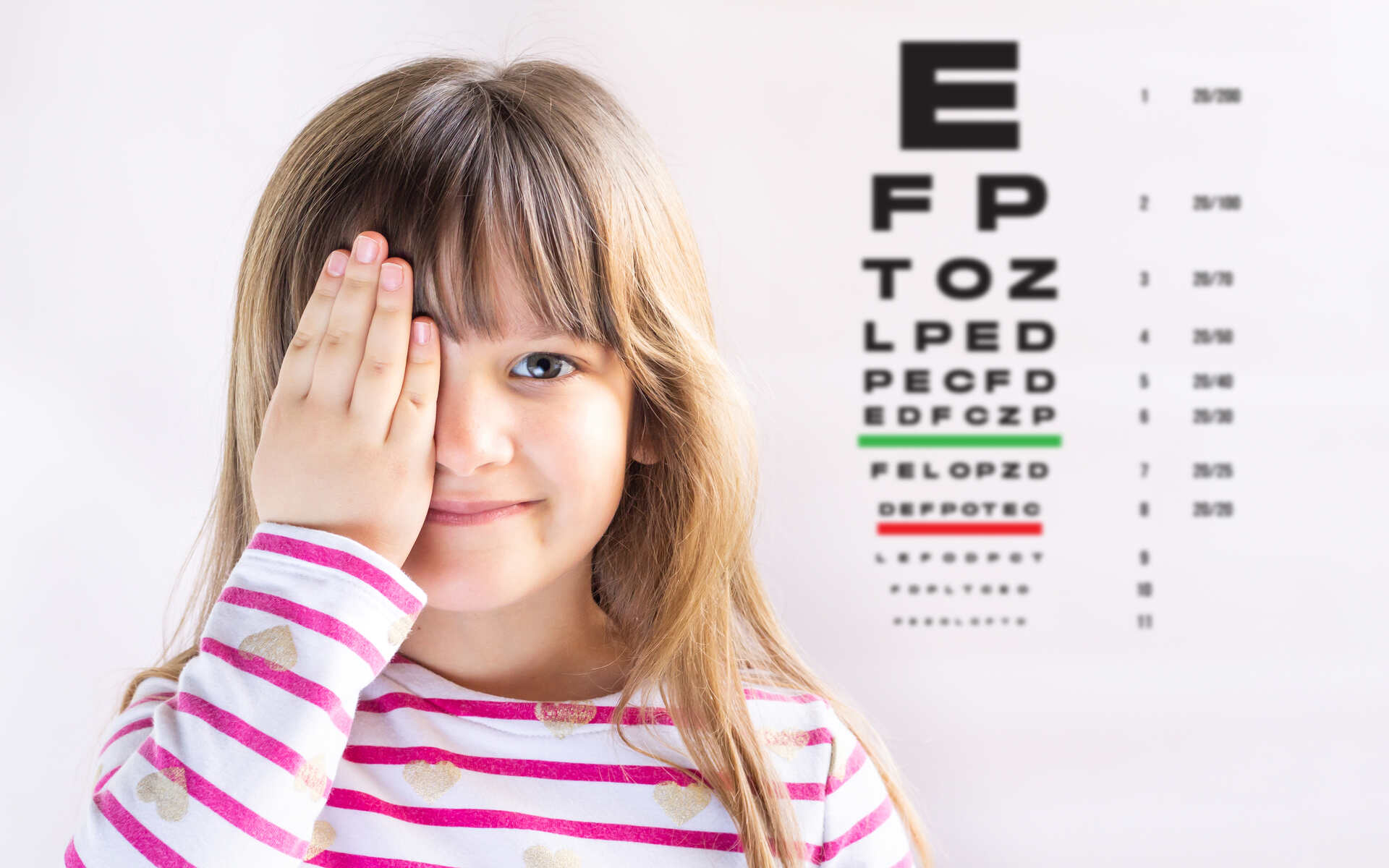 ok.vision Children’s Eye Care A young girl covering one eye while smiling, standing in front of an eye chart for Children’s Eye Care.