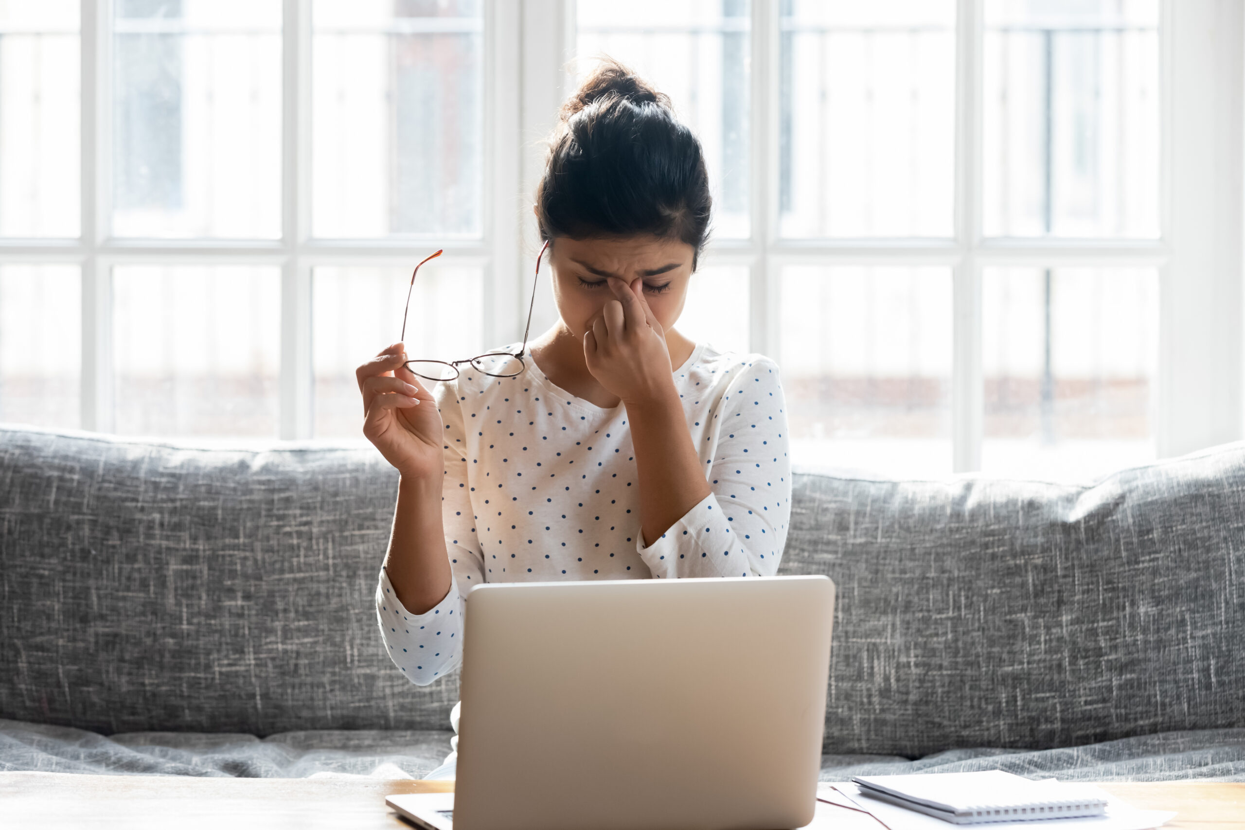 Tired,Young,Woman,Take,Off,Glasses,Massage,Eyes,Overwhelmed,With A woman sitting on a couch, rubbing her eyes while holding her glasses, highlighting concerns about eye health and digital strain.