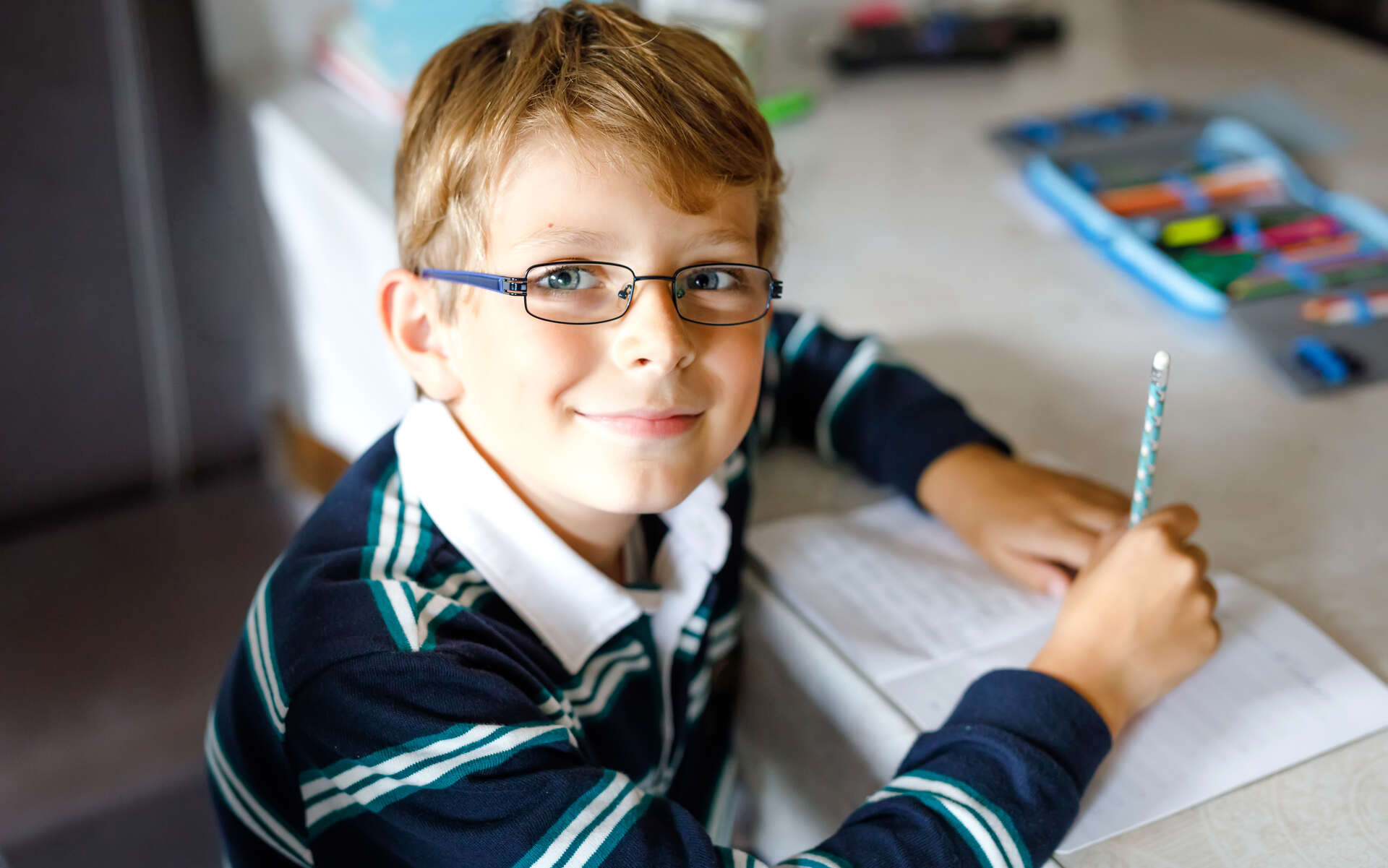 A young boy wearing glasses smiles while writing in a notebook, highlighting the importance of eye health exercises during classroom activities.