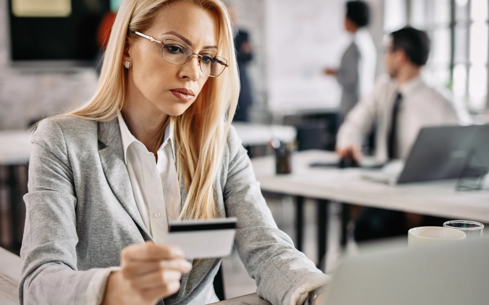 woman-paying-with-credit-card A professional woman in an office holds a credit card while looking at her laptop, symbolizing shifting benefits to employees through flexible payment or reimbursement systems.
